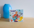 Photo du coffret 3 CDs de Jean René posé sur une table en bois, montrant l'illustration de couverture avec guitare et objets flottants.